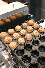 A kitchen appliance with a non-stick mold is used to cook small, round pastries. The pastries are light brown and appear to be baked evenly. The appliance has a metal lid and several of the molds are empty, giving a glimpse of the cooking surface.