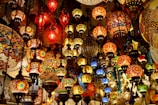An array of colorful, intricately designed mosaic lamps hanging closely together. The lamps vary in size and shape, casting a warm, inviting glow with vibrant shades of red, orange, yellow, blue, and green. The patterns and detailed craftsmanship make each lamp unique, creating a lively and exotic atmosphere.