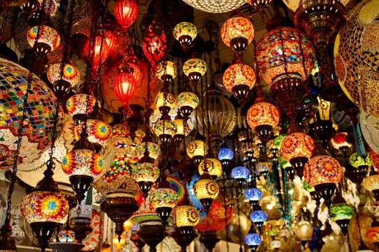 An array of colorful, intricately designed mosaic lamps hanging closely together. The lamps vary in size and shape, casting a warm, inviting glow with vibrant shades of red, orange, yellow, blue, and green. The patterns and detailed craftsmanship make each lamp unique, creating a lively and exotic atmosphere.