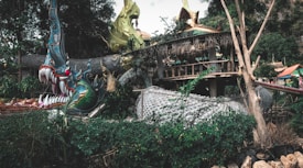 A large, intricately designed dragon sculpture intertwines with surrounding foliage and structures. The dragon features vibrant green, blue, and gold hues with sharp teeth and intense red eyes, giving it a fierce and mystical appearance. The background includes a rustic, thatched roof structure surrounded by lush greenery.
