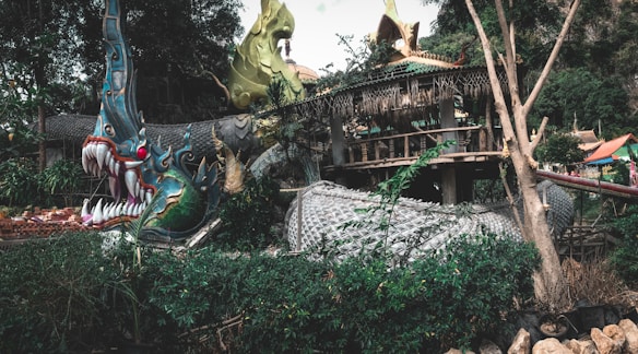 A large, intricately designed dragon sculpture intertwines with surrounding foliage and structures. The dragon features vibrant green, blue, and gold hues with sharp teeth and intense red eyes, giving it a fierce and mystical appearance. The background includes a rustic, thatched roof structure surrounded by lush greenery.