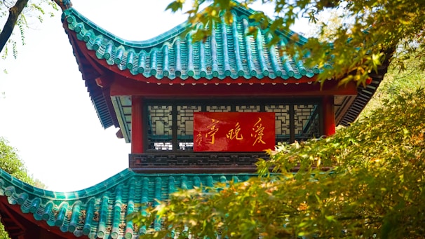 A traditional Asian pavilion with green and turquoise tiled roof, partially obscured by lush green foliage. A red sign with gold script is prominently displayed on the building's facade.