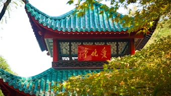 A traditional Asian pavilion with green and turquoise tiled roof, partially obscured by lush green foliage. A red sign with gold script is prominently displayed on the building's facade.