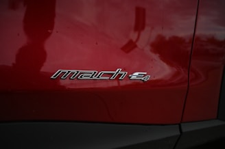 A close-up of a red vehicle's side panel featuring a silver emblem with the text 'mach e4'. The surface is glossy, reflecting the surrounding environment faintly.
