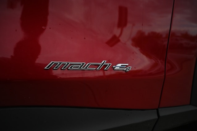A close-up of a red vehicle's side panel featuring a silver emblem with the text 'mach e4'. The surface is glossy, reflecting the surrounding environment faintly.