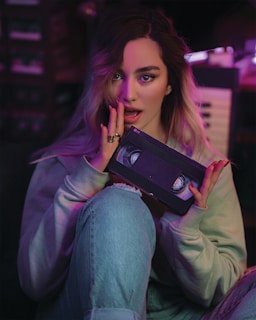 A person with long hair is holding a VHS tape in a dimly lit room. Soft purple and pink lighting casts a retro ambiance. They are wearing a light-colored sweater and jeans while seated.