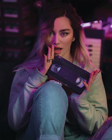 A person with long hair is holding a VHS tape in a dimly lit room. Soft purple and pink lighting casts a retro ambiance. They are wearing a light-colored sweater and jeans while seated.