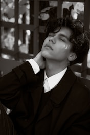 A person is leaning back with eyes closed, dressed in a formal collared shirt and dark jacket. The pose conveys a sense of relaxation or introspection. The background includes a pattern created by light and shadow against a textured surface.