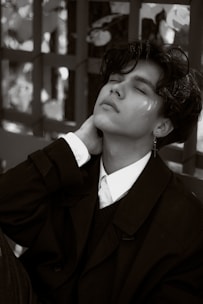 A person is leaning back with eyes closed, dressed in a formal collared shirt and dark jacket. The pose conveys a sense of relaxation or introspection. The background includes a pattern created by light and shadow against a textured surface.