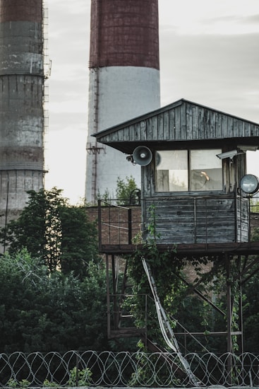 A rustic wooden watchtower with metal railings stands amidst dense greenery. Vines climb up the structure, which has two loudspeakers attached. In the background, there are large, industrial smokestacks with red and white sections.