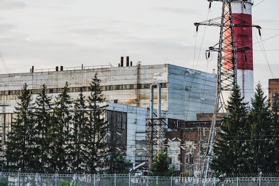 An industrial complex with large factory buildings, a tall red and white striped chimney, and metal power lines in the foreground. Tall evergreen trees partially obscure some of the structures.