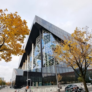 A modern architectural building with a striking glass facade and angular design is set against a backdrop of autumn trees with golden leaves. The contemporary structure features a complex pattern of intersecting lines and reflective surfaces. Around the building, bicycles are parked and there are a few street signs visible, indicating an urban setting.