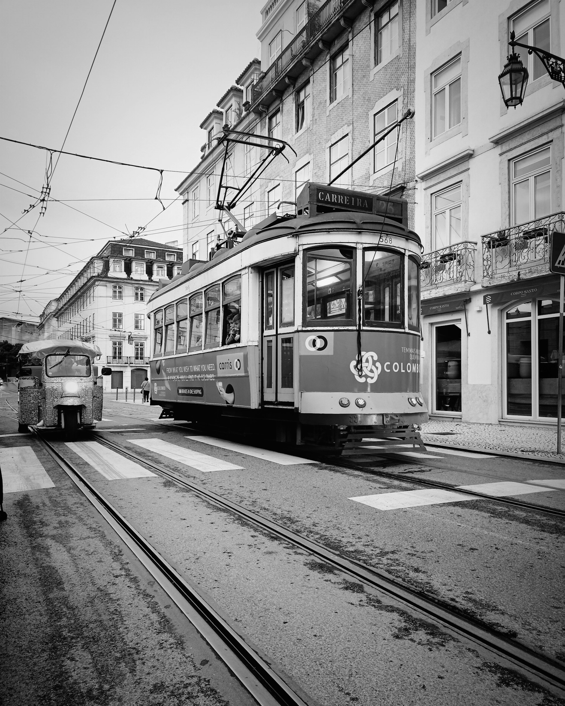 Square black and white image showing a vintage tram crossing a bustling Genoa street, framed by historic buildings and dynamic light contrasts.