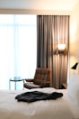 A well-lit hotel room with a large window covered by sheer white curtains and grey drapes. A comfortable brown armchair sits next to a small round glass table, accompanied by a tall floor lamp with a light shade. In the foreground, an unmade bed with white linens has a dark shirt casually lying on top of it.