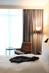 A well-lit hotel room with a large window covered by sheer white curtains and grey drapes. A comfortable brown armchair sits next to a small round glass table, accompanied by a tall floor lamp with a light shade. In the foreground, an unmade bed with white linens has a dark shirt casually lying on top of it.
