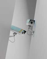 Two surveillance cameras are mounted on a stark, minimalist wall. The cameras have a teal body with orange accents and are connected by black cables. They are angled in different directions, suggesting active observation.