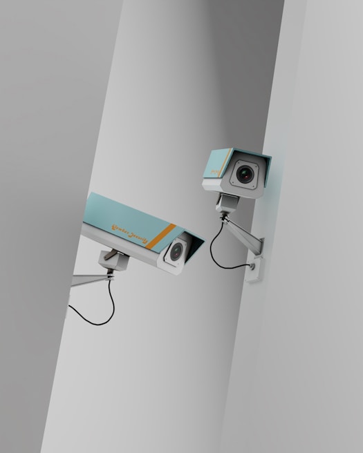 Two surveillance cameras are mounted on a stark, minimalist wall. The cameras have a teal body with orange accents and are connected by black cables. They are angled in different directions, suggesting active observation.