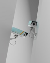 Two surveillance cameras are mounted on a stark, minimalist wall. The cameras have a teal body with orange accents and are connected by black cables. They are angled in different directions, suggesting active observation.