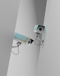 Two surveillance cameras are mounted on a stark, minimalist wall. The cameras have a teal body with orange accents and are connected by black cables. They are angled in different directions, suggesting active observation.