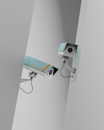 Two surveillance cameras are mounted on a stark, minimalist wall. The cameras have a teal body with orange accents and are connected by black cables. They are angled in different directions, suggesting active observation.