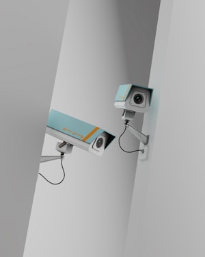Two surveillance cameras are mounted on a stark, minimalist wall. The cameras have a teal body with orange accents and are connected by black cables. They are angled in different directions, suggesting active observation.