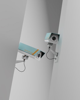 Two surveillance cameras are mounted on a stark, minimalist wall. The cameras have a teal body with orange accents and are connected by black cables. They are angled in different directions, suggesting active observation.