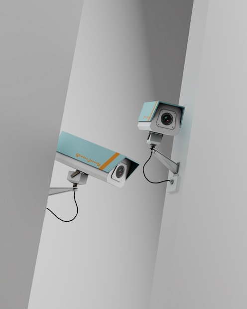Two surveillance cameras are mounted on a stark, minimalist wall. The cameras have a teal body with orange accents and are connected by black cables. They are angled in different directions, suggesting active observation.