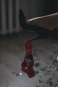 A leg in a black stiletto heel is shown with a red festive thong featuring a snowman design hanging from the shoe. The floor is scattered with multicolored confetti, suggesting a playful or celebratory setting.