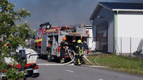 Firefighters in protective gear are operating near a fire truck, handling hoses. The scene is set on a road adjacent to a building, with smoke visible in the background. A fence and a few trees are also present.
