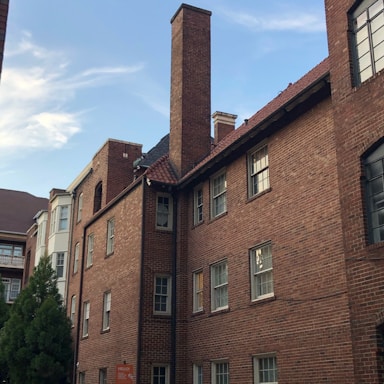 A multi-story red brick building with chimneys, rectangular windows, and a sloped roof is located in an urban setting. The building is adjacent to another structure with a mix of architectural styles. A tree with dense foliage is visible in the foreground.