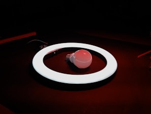 A glowing white circular ring light encircles a single incandescent light bulb lying on a dark surface. The ambient lighting has a warm red tint, creating a contrast with the bright white of the ring light.