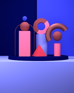 A collection of geometric shapes is arranged on a stage with a dark blue backdrop. The setup includes a pink cylinder topped with a brown sphere, a pink and brown donut shape, a blue cylinder, a pink triangular pyramid, and a pink cylinder topped with a brown arc. The overall composition is minimalistic yet vibrant, with a strong contrast between the shapes and their background.