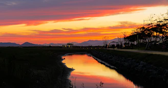 A vibrant orange-hued sunset over the serene Yamuna river in Vrindavan, with pilgrims walking towards a distant temple.