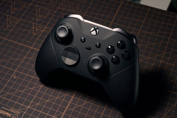 A sleek black gaming controller resting on a brown grid-patterned cutting mat. The controller has textured grips, two analog sticks, a directional pad, and several buttons including X, Y, B, and A. The lighting casts a subtle shadow, highlighting its ergonomic design.