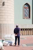 A person wearing a cap stands on a paved area with their back turned, holding a piece of paper behind their back. Large bags are stacked nearby, and the person is facing the wall of a building with a distinct arched window and light fixture.