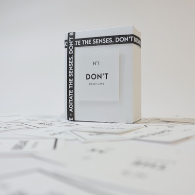 A minimalist perfume packaging design features a white box with black text. The packaging has a sleek, modern aesthetic with the words 'N°1 DON’T PERFUME' displayed prominently on the front. A black strap with white text wraps around the box, adding contrast and interest to the design.