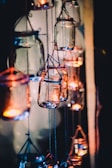 Multiple glass jars hang in a dimly lit setting, each containing a small candle that emits a warm, glowing light. The jars are suspended by blue twine, creating a whimsical and cozy atmosphere.