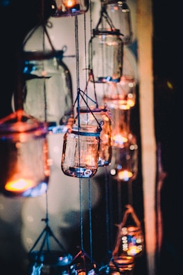 Multiple glass jars hang in a dimly lit setting, each containing a small candle that emits a warm, glowing light. The jars are suspended by blue twine, creating a whimsical and cozy atmosphere.