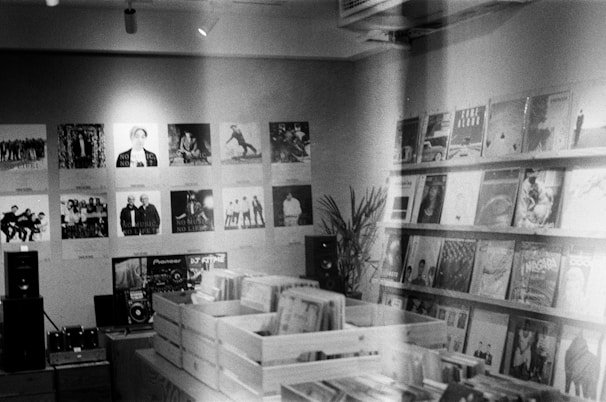 A vintage-style record store with walls adorned with music posters. Wooden crates in the center hold vinyl records, and shelves display various album covers. A small sound system setup is visible on a table, and a plant adds a touch of greenery to the scene.