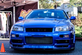 A vibrant blue sports car is parked outdoors with various street elements around it. A clothing rack with hanging garments is visible to the left, accompanied by a pop-up tent. An orange traffic cone is positioned near the front of the car.