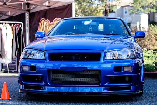A vibrant blue sports car is parked outdoors with various street elements around it. A clothing rack with hanging garments is visible to the left, accompanied by a pop-up tent. An orange traffic cone is positioned near the front of the car.