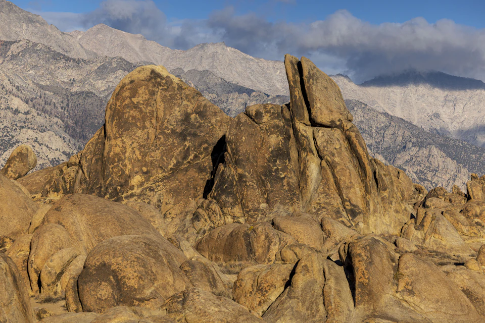 The image depicts a rocky landscape with large boulders scattered across the scene, some of which have been weathered by time and elements. The boulders vary in size and shape, creating a sense of depth and dimension to the landscape. In the background, majestic mountains rise against the sky, their peaks touching the clouds. The colors in the image are predominantly browns and grays, with patches of blue from the sky and clouds adding contrast.