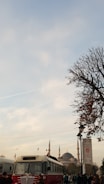 A vintage red and white bus is parked on a street with a historical mosque with tall minarets in the background. There are a few people walking around, and a tree with sparse leaves is on the right side of the image. The sky is partly cloudy with contrails visible.