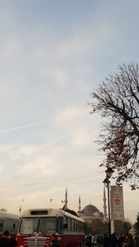 A vintage red and white bus is parked on a street with a historical mosque with tall minarets in the background. There are a few people walking around, and a tree with sparse leaves is on the right side of the image. The sky is partly cloudy with contrails visible.