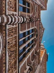 A detailed architectural facade featuring intricate carvings and sculptures. The surface displays a pattern of dark bricks interspersed with lighter stone elements. Several tall, narrow windows with reflective glass are framed by the structure, adding elegance. Ornamental sculptures and decorative reliefs are integrated into the building’s design.