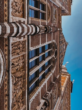 A detailed architectural facade featuring intricate carvings and sculptures. The surface displays a pattern of dark bricks interspersed with lighter stone elements. Several tall, narrow windows with reflective glass are framed by the structure, adding elegance. Ornamental sculptures and decorative reliefs are integrated into the building’s design.