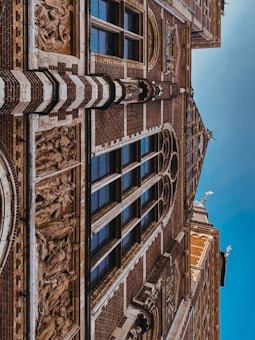 A detailed architectural facade featuring intricate carvings and sculptures. The surface displays a pattern of dark bricks interspersed with lighter stone elements. Several tall, narrow windows with reflective glass are framed by the structure, adding elegance. Ornamental sculptures and decorative reliefs are integrated into the building’s design.