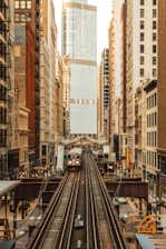 An urban scene featuring a train on elevated tracks moving through a cityscape. Tall buildings line both sides of the tracks, with a prominent skyscraper in the center displaying the name 'TRUMP'. The architecture varies from older brick buildings to modern glass structures.