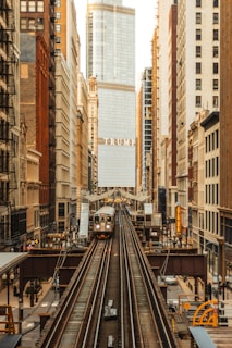 An urban scene featuring a train on elevated tracks moving through a cityscape. Tall buildings line both sides of the tracks, with a prominent skyscraper in the center displaying the name 'TRUMP'. The architecture varies from older brick buildings to modern glass structures.