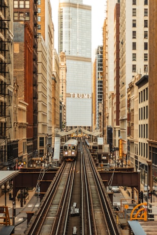An urban scene featuring a train on elevated tracks moving through a cityscape. Tall buildings line both sides of the tracks, with a prominent skyscraper in the center displaying the name 'TRUMP'. The architecture varies from older brick buildings to modern glass structures.